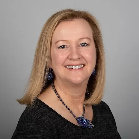 A woman with shoulder-length blonde hair, wearing a black top, blue jewelry, and smiling at the camera against a gray background.