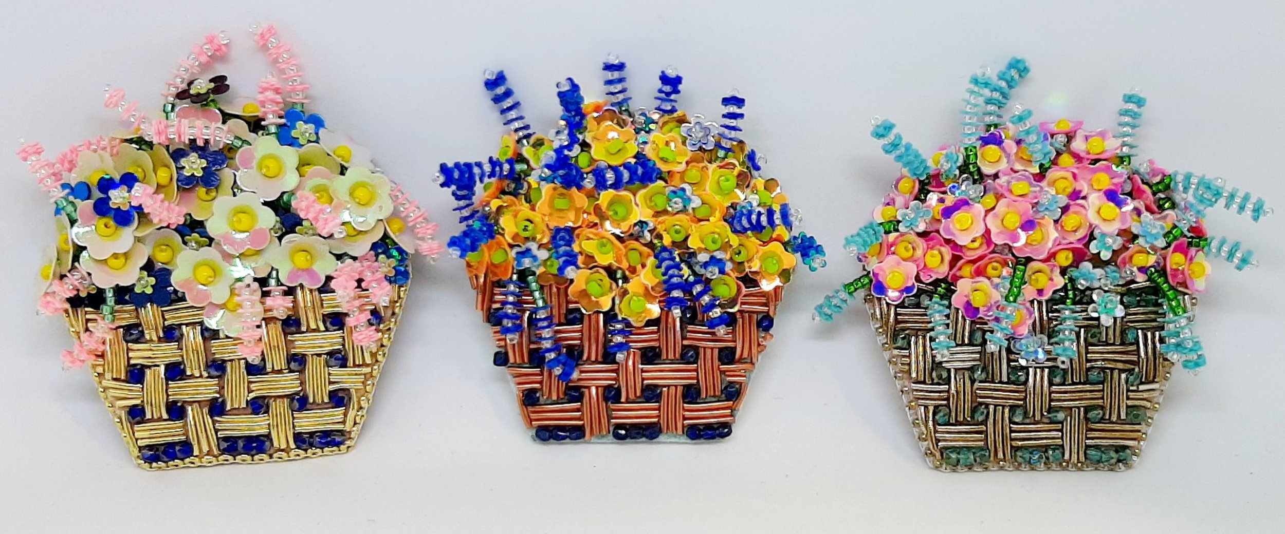 Three colorful beaded flower baskets. Each basket is decorated with miniature beaded flowers and greenery, in pink, blue, and teal color schemes, arranged on a white background.