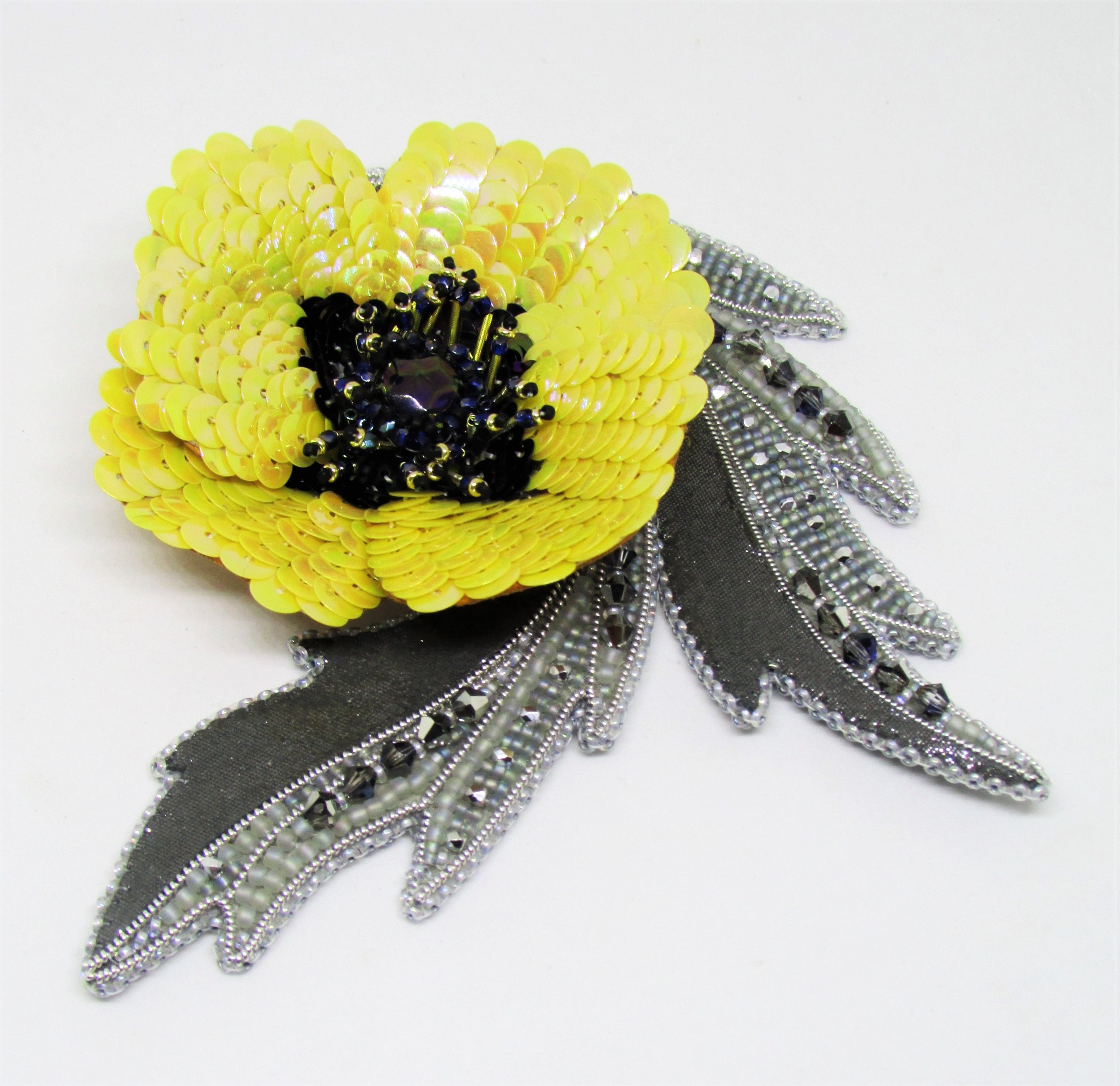 A yellow flower-shaped brooch made of sequins with black and purple accents and a silver mesh ribbon beneath it.