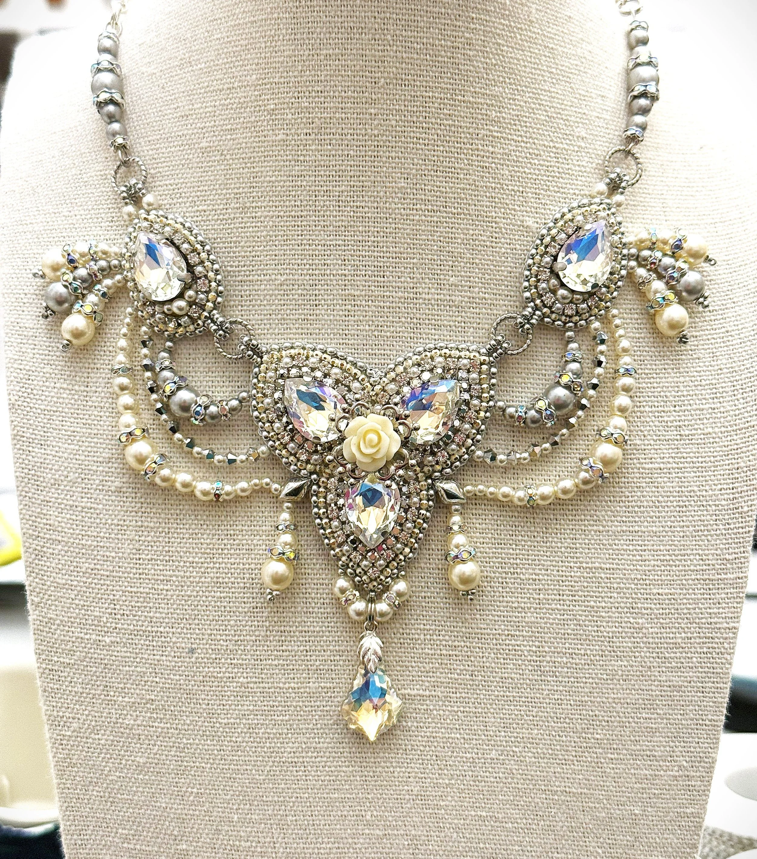 Elegant statement necklace with large teardrop-shaped crystals, surrounded by smaller rhinestones, pearls, and floral accents, displayed on a beige fabric bust.