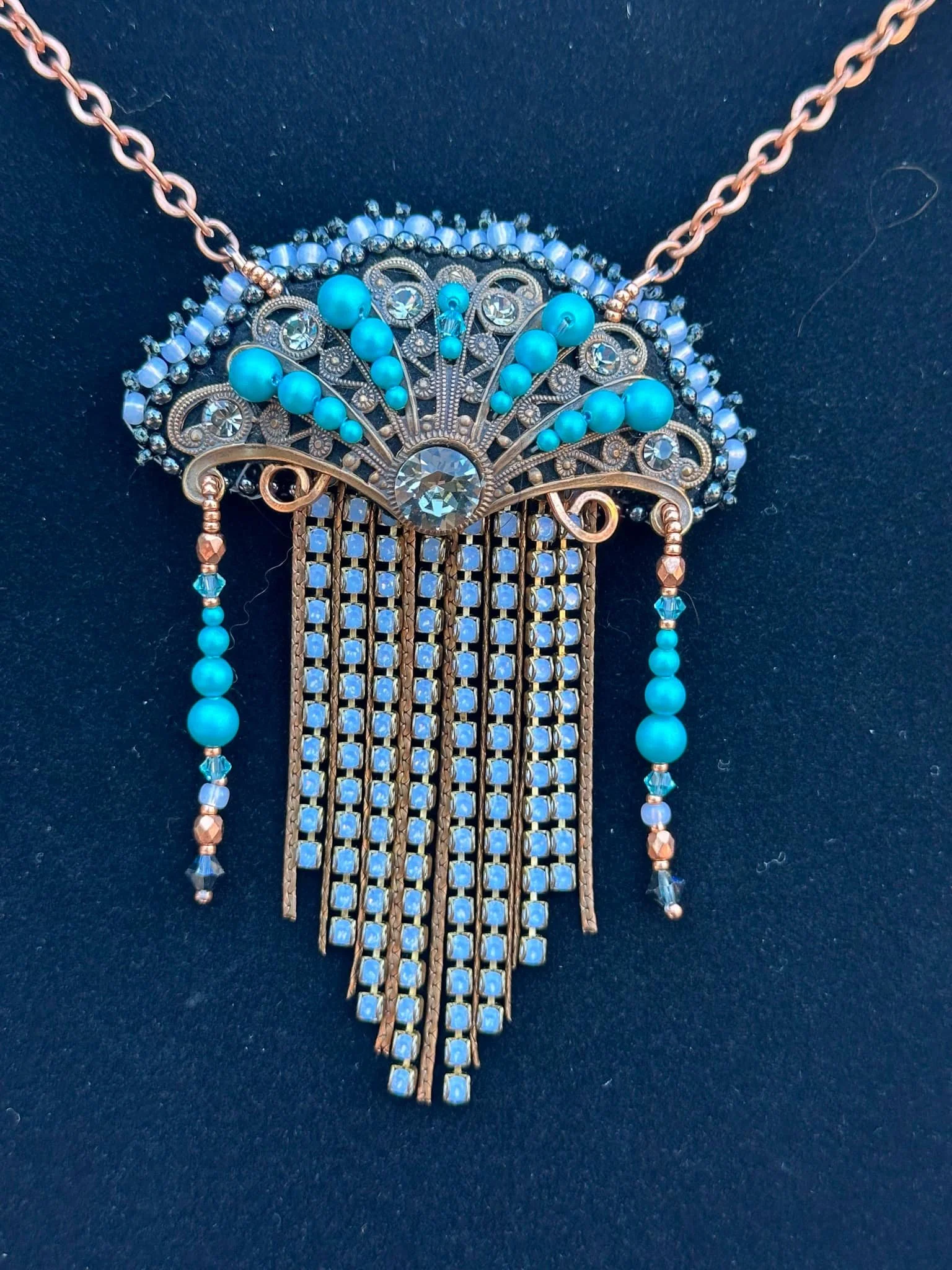 A decorative necklace with intricate gold metalwork, blue beads, and clear rhinestones, featuring dangling bead strands and a large central rhinestone.