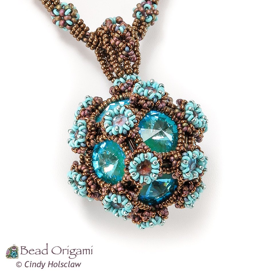 Beaded jewelry pendant with brown, turquoise, and pink beads surrounding blue stones in a complex, woven design.