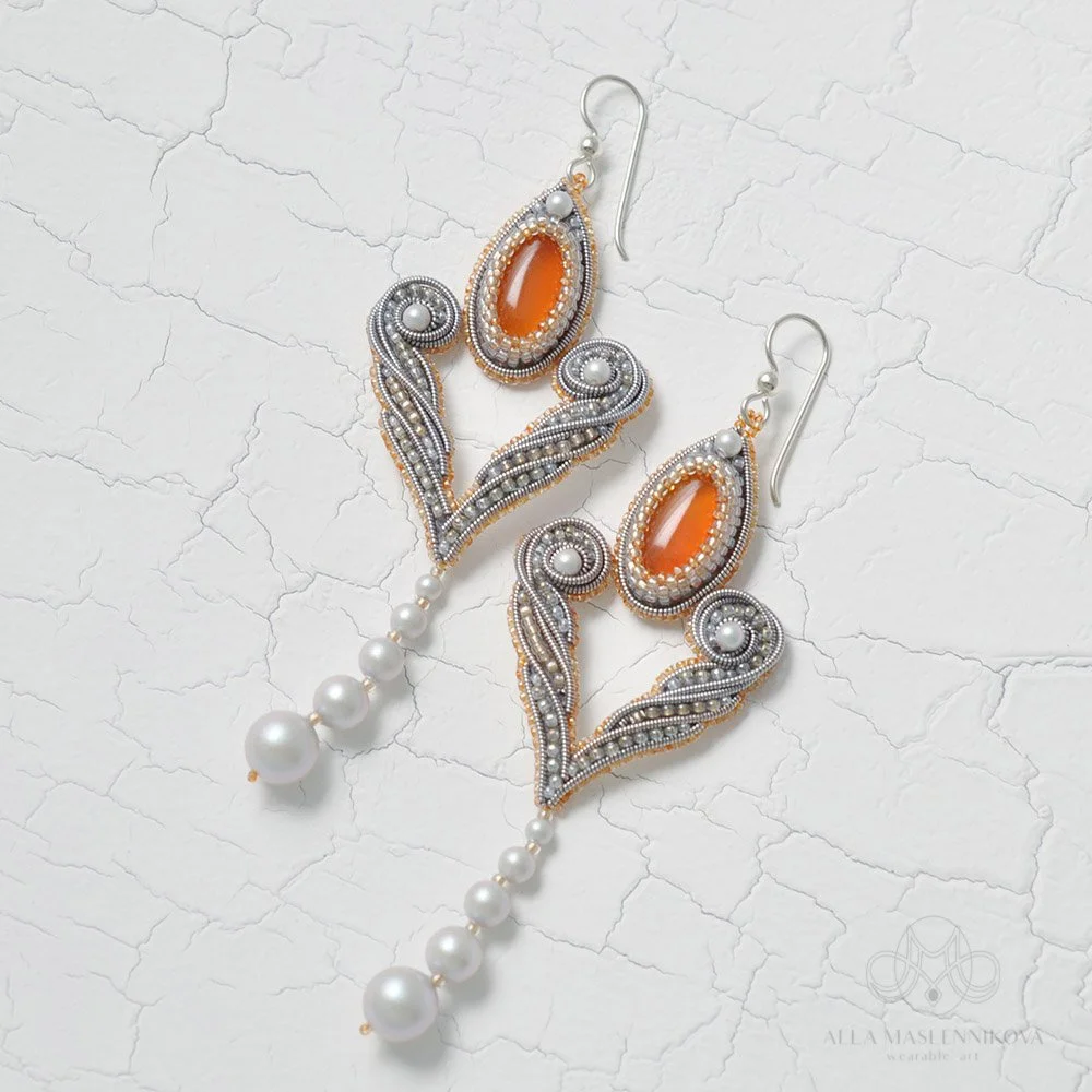 Pair of intricate earrings with orange oval stones, detailed beadwork, silver and gold accents, and hanging pearls, set on a white textured surface.