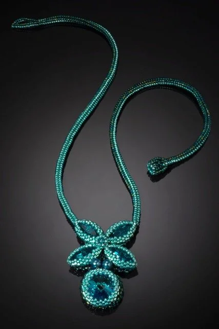 A turquoise-colored rhinestone necklace with a circular pendant and a bow-shaped design in the center, displayed on a black background.