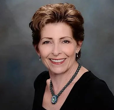 A smiling woman with short brown hair, wearing a black top and a beaded necklace, against a dark gray background.