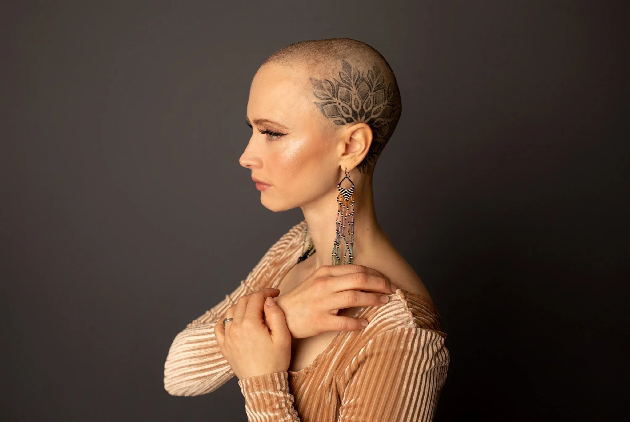 A woman with a shaved head and a large floral tattoo on the side of her head, wearing earrings and a striped top, standing against a dark background.
