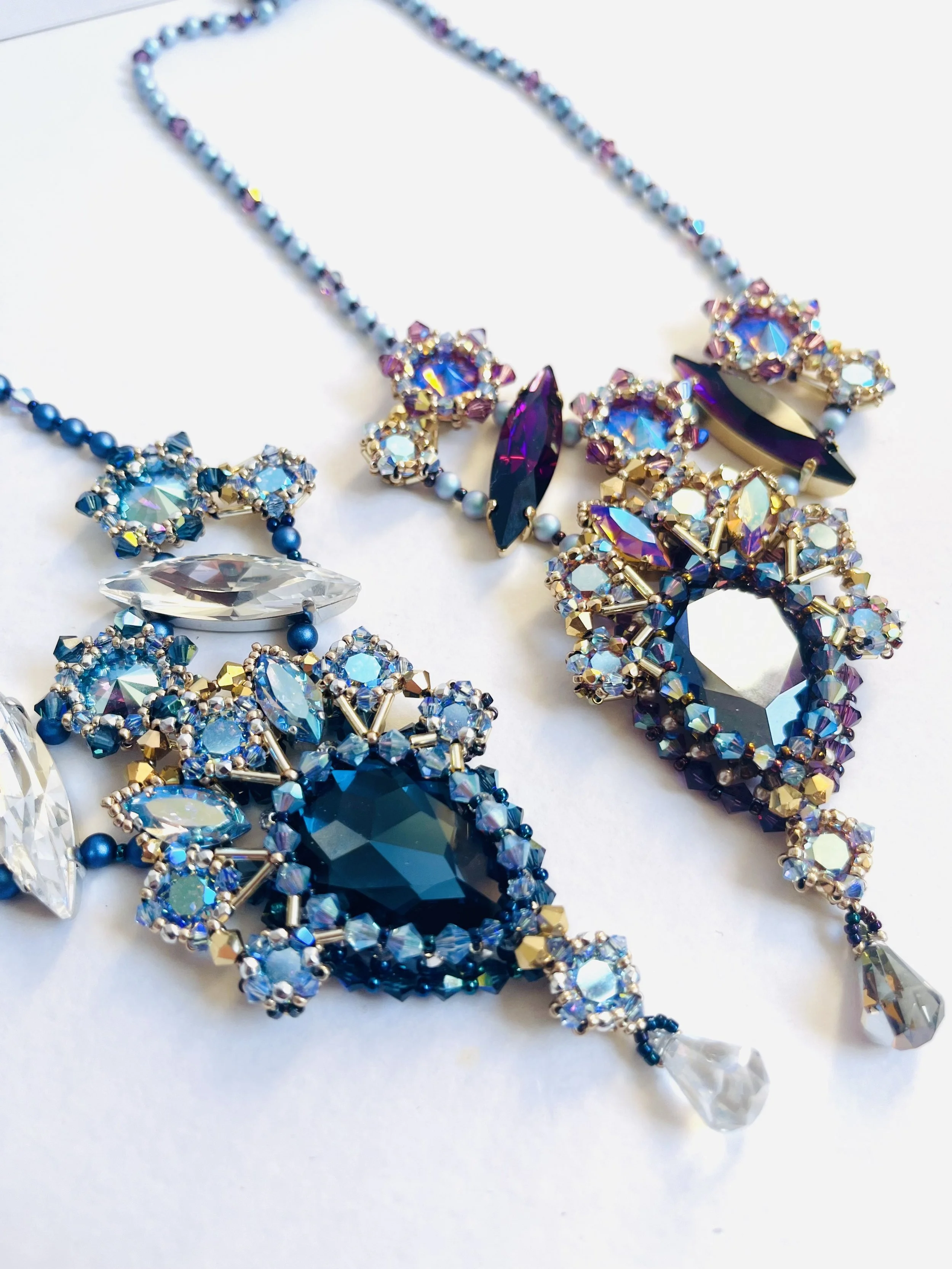 Close-up of a colorful beaded jewelry set with blue, purple, gold, and clear rhinestones, featuring necklaces and earrings arranged on a white surface.