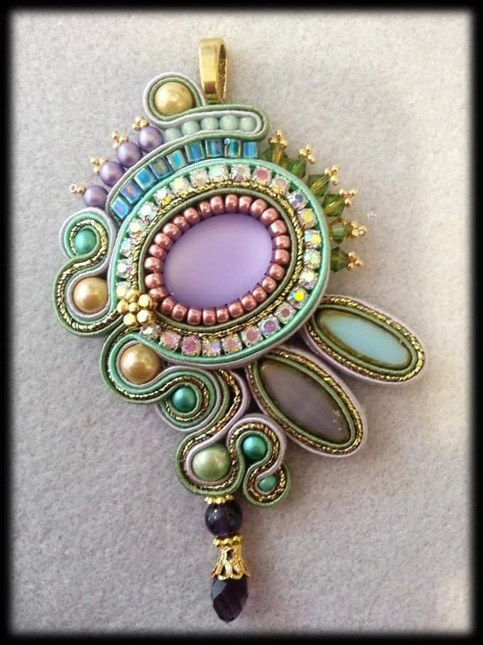 A colorful ornate pendant with beads, rhinestones, and intricate fabric or thread work in pastel shades of purple, green, pink, and gold.