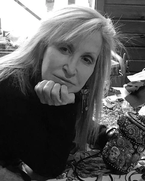 A woman with blonde hair resting her chin on her hand, looking at the camera in a dimly lit indoor setting with cluttered items around her.