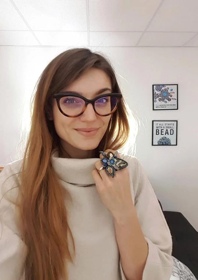 A woman with long red hair, wearing glasses and a cream turtleneck sweater, smiling while holding her hand near her face with a large, decorative butterfly-shaped ring.