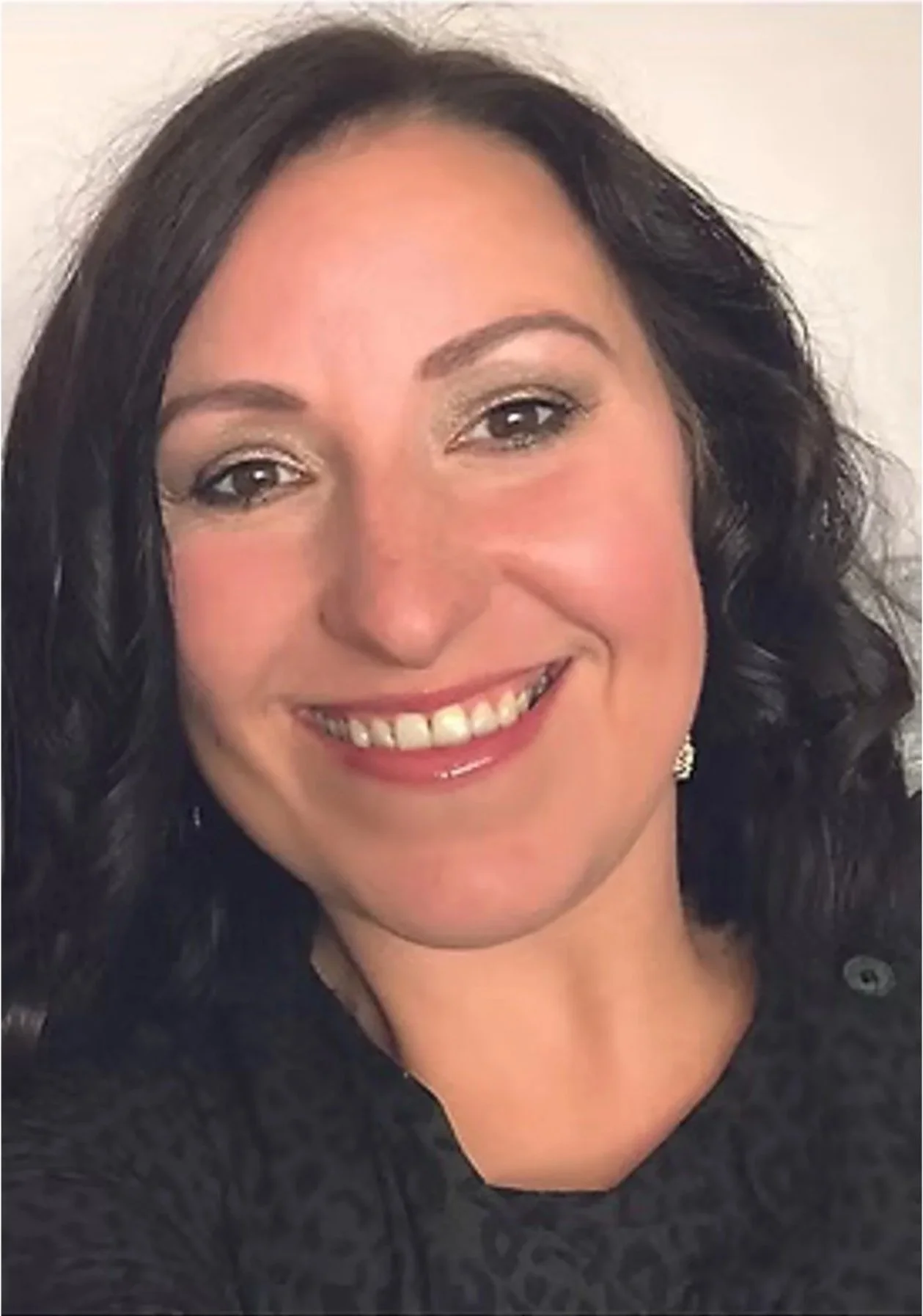 A woman with dark hair styled in curls, smiling, wearing a black top and earrings, against a plain light background.