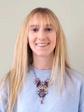 A woman with long blonde hair, wearing a light blue sweater and a large ornate necklace, smiles at the camera.