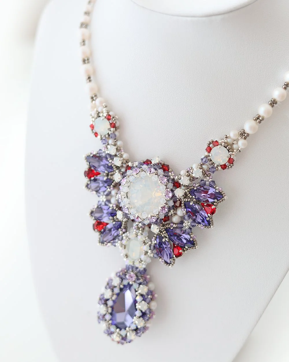Close-up of a colorful statement necklace with purple, red, white, and clear gemstones, displayed on a white bust.