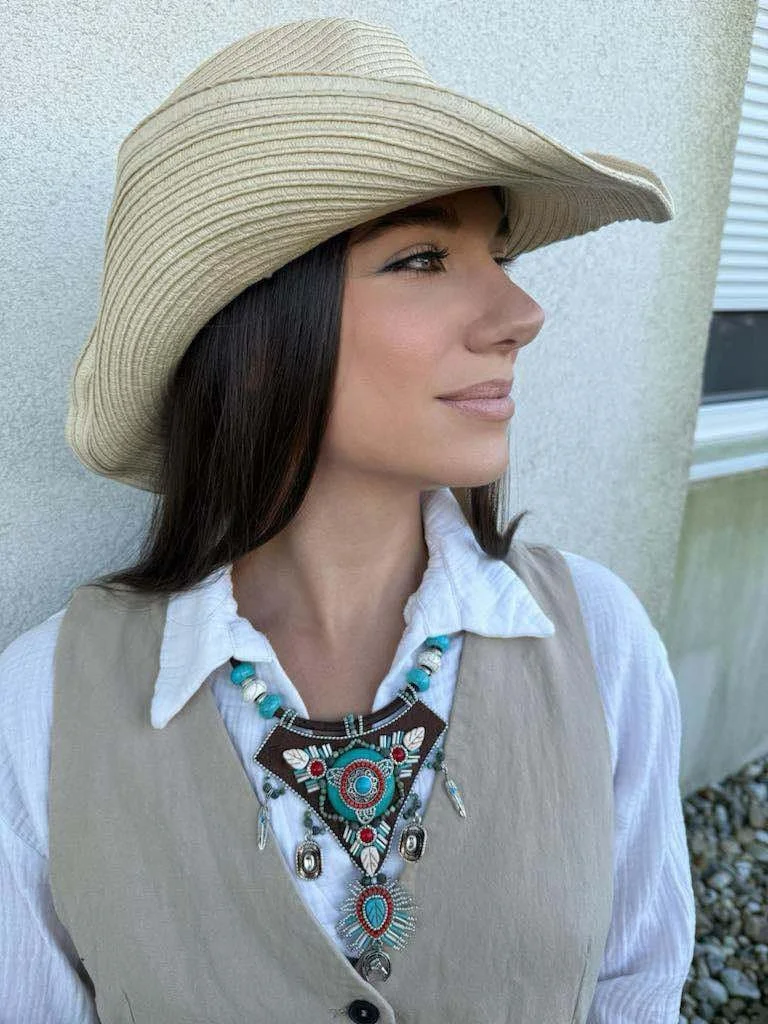 Profile of a woman wearing a wide-brimmed straw hat, white shirt, beige vest, and a layered turquoise and silver necklace, standing outdoors against a light-colored wall.