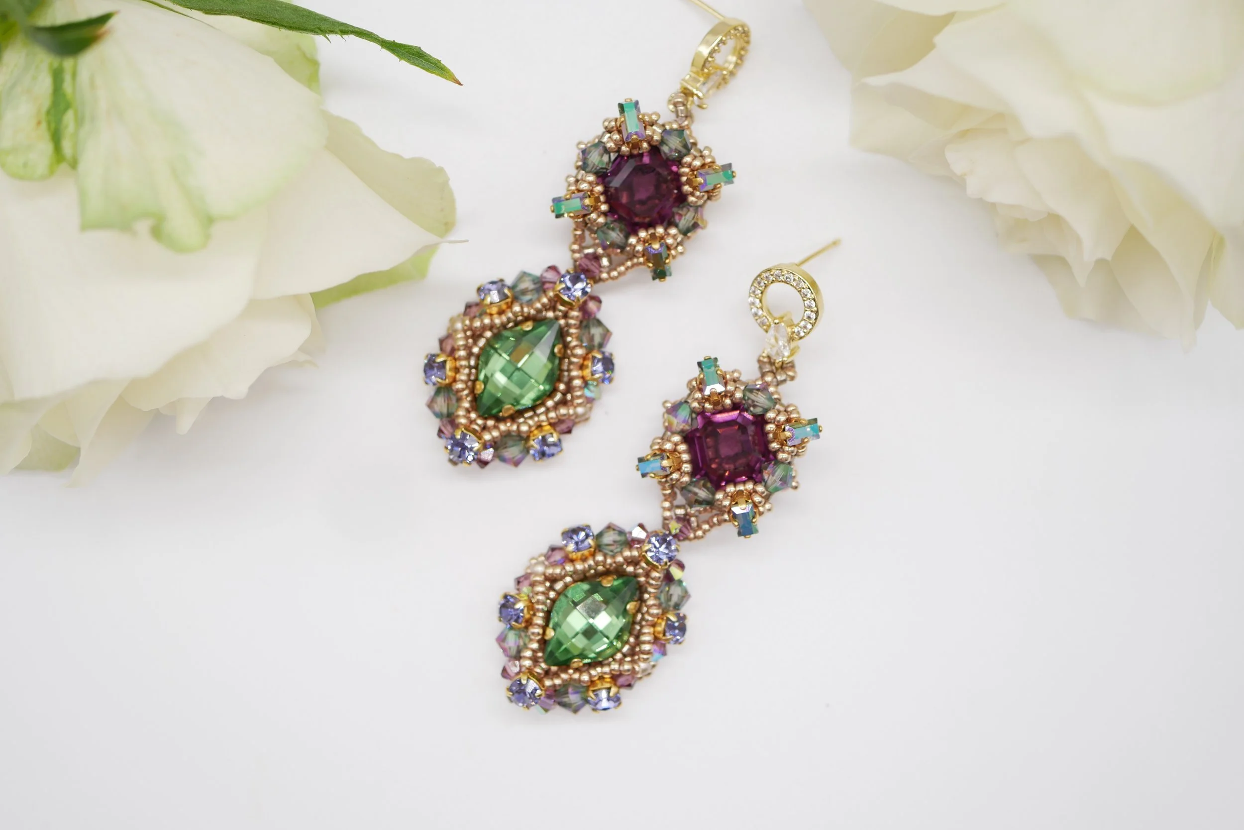 Jewelry set with large green and purple gemstones, gold accents, and surrounding smaller crystals, displayed on a white background with white flowers.