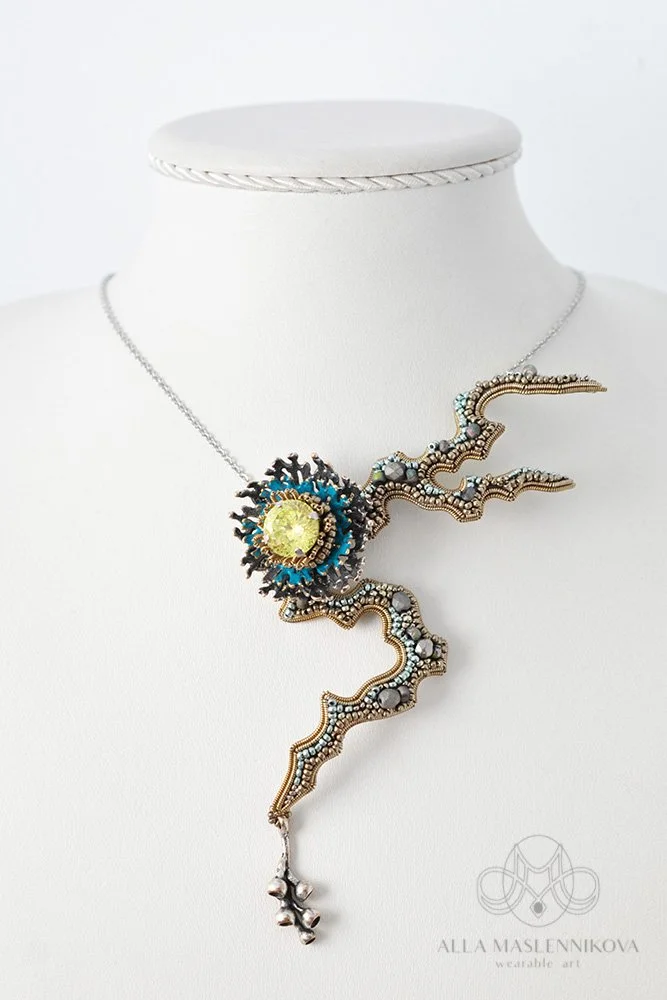 Elegant necklace with a large yellow gemstone at the center, resembling a sun, attached to a wavy, intricate metal chain with bead details, displayed on a white bust.