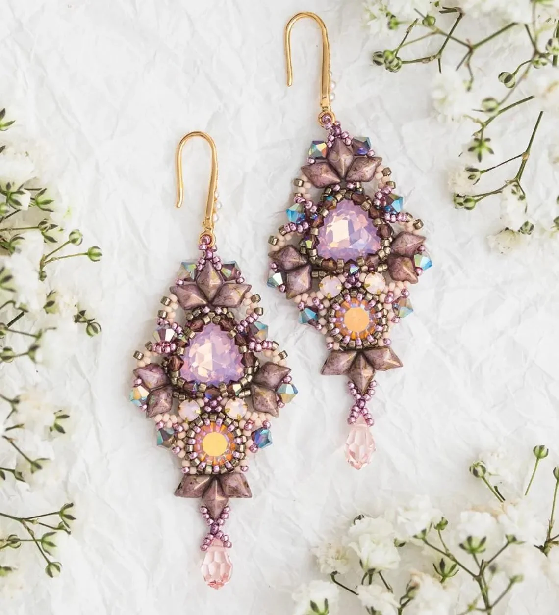 Pair of ornate purple and pink beaded earrings with large central crystals, surrounded by smaller beads and flower-shaped accents, hanging on crinkled white background with white flowers nearby.
