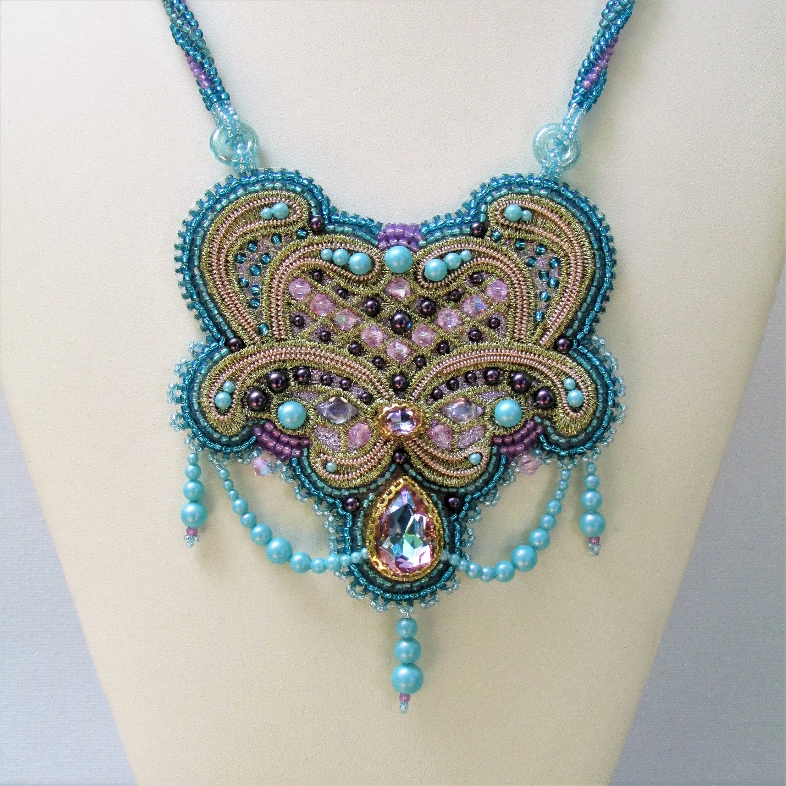 Close-up of a colorful, beaded necklace with intricate designs, featuring beads in shades of blue, pink, purple, and gold, displayed on a white mannequin.