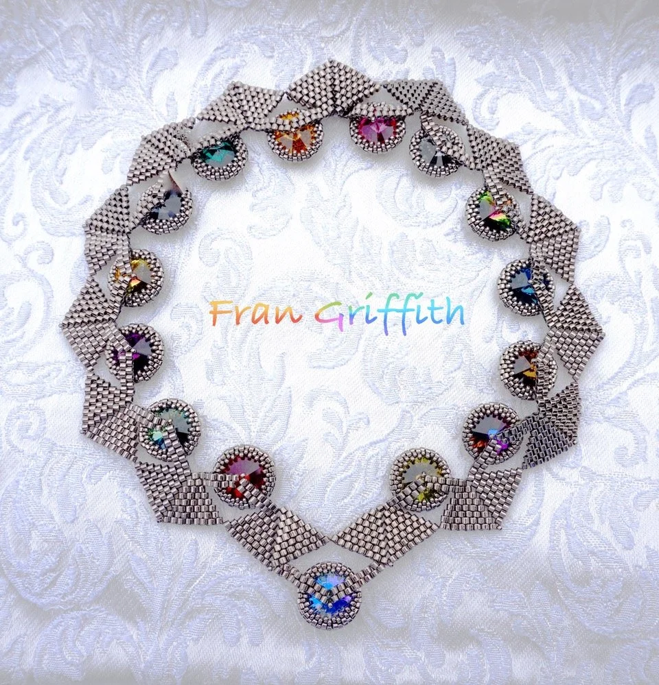 A silver bracelet with multicolored gemstones arranged in a circular pattern, placed on a white textured fabric background. The words "Fran Griffith" are written in rainbow-colored text in the center.