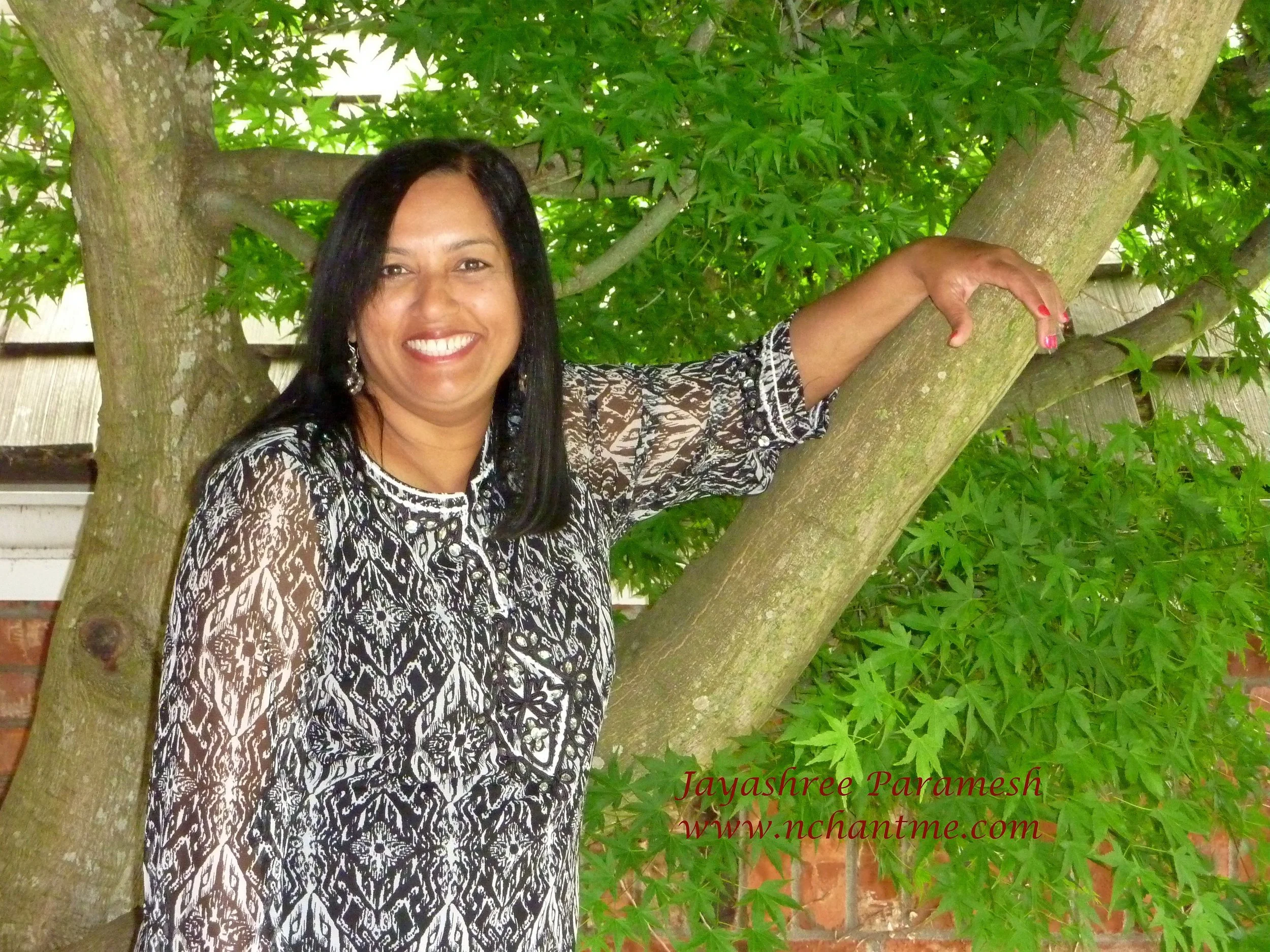 Woman with long black hair and a big smile, wearing a black and white patterned top, standing in front of a tree with green leaves and a brick wall in the background.