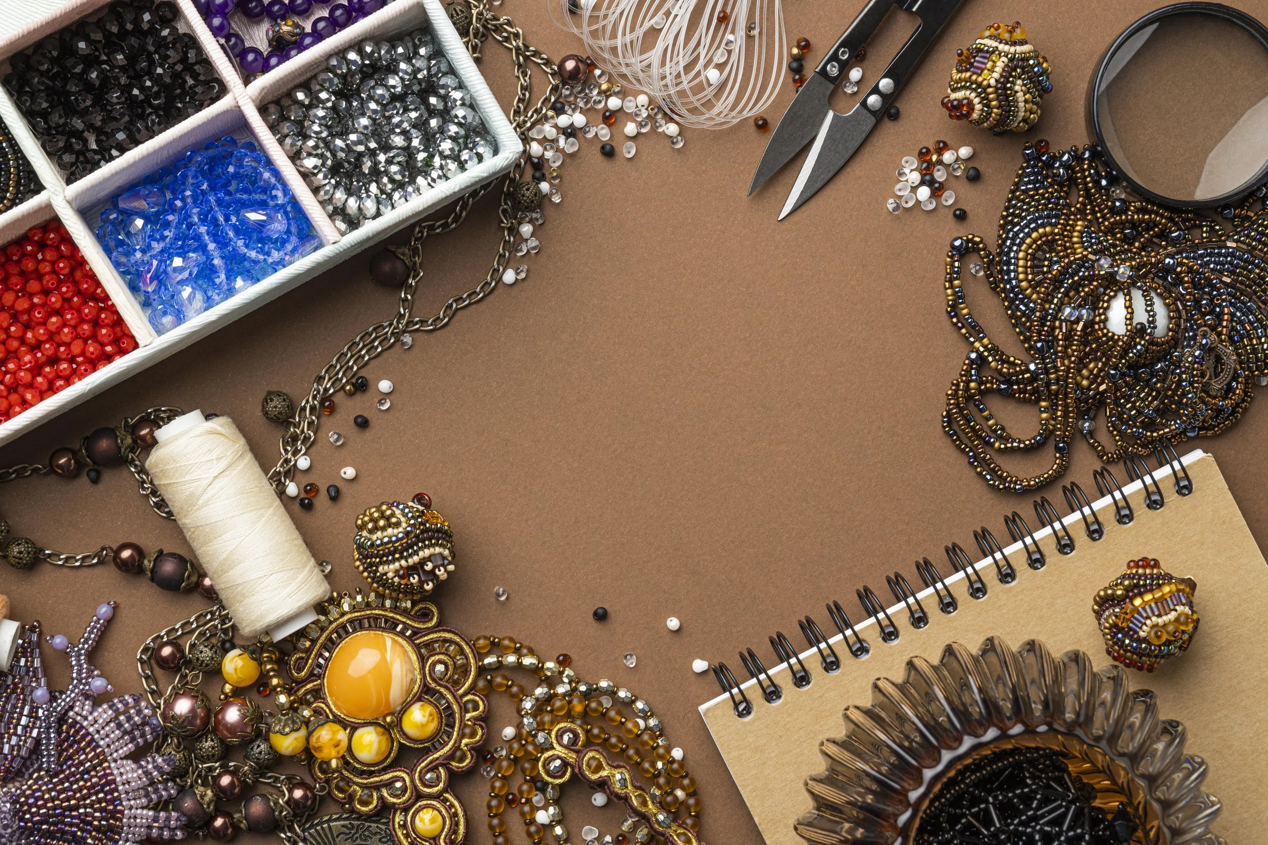 A variety of jewelry making supplies, including beads, chains, scissors, and a notebook, arranged on a brown surface.