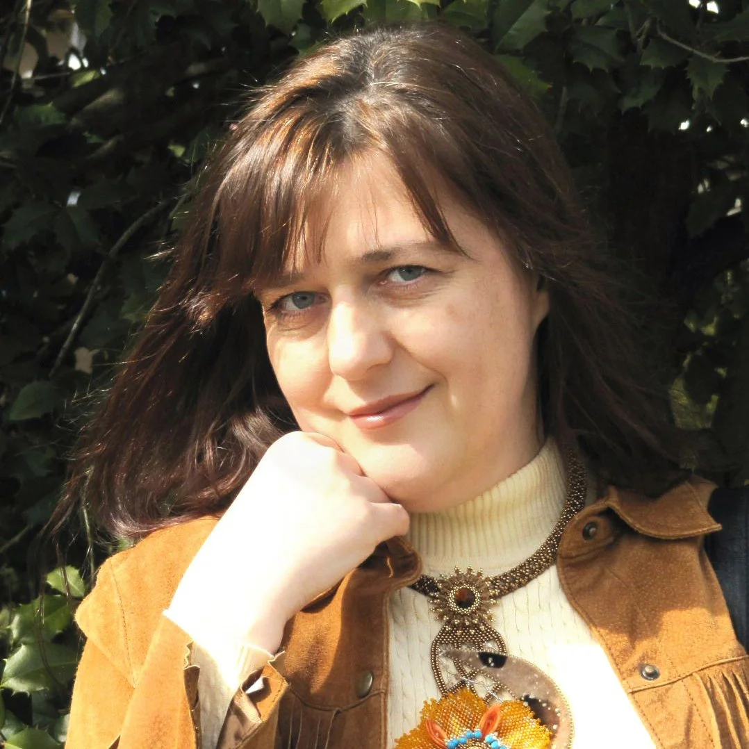 A woman with shoulder-length brown hair and blue eyes smiling outdoors near leafy background.