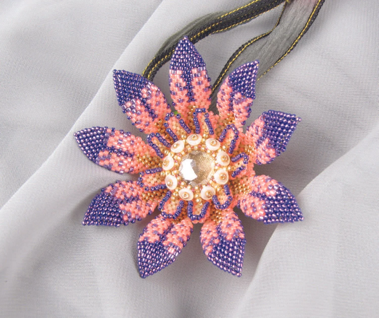 Beaded floral necklace with purple, pink, and gold beads, and a central crystal embellishment, resting on a gray and white fabric background.