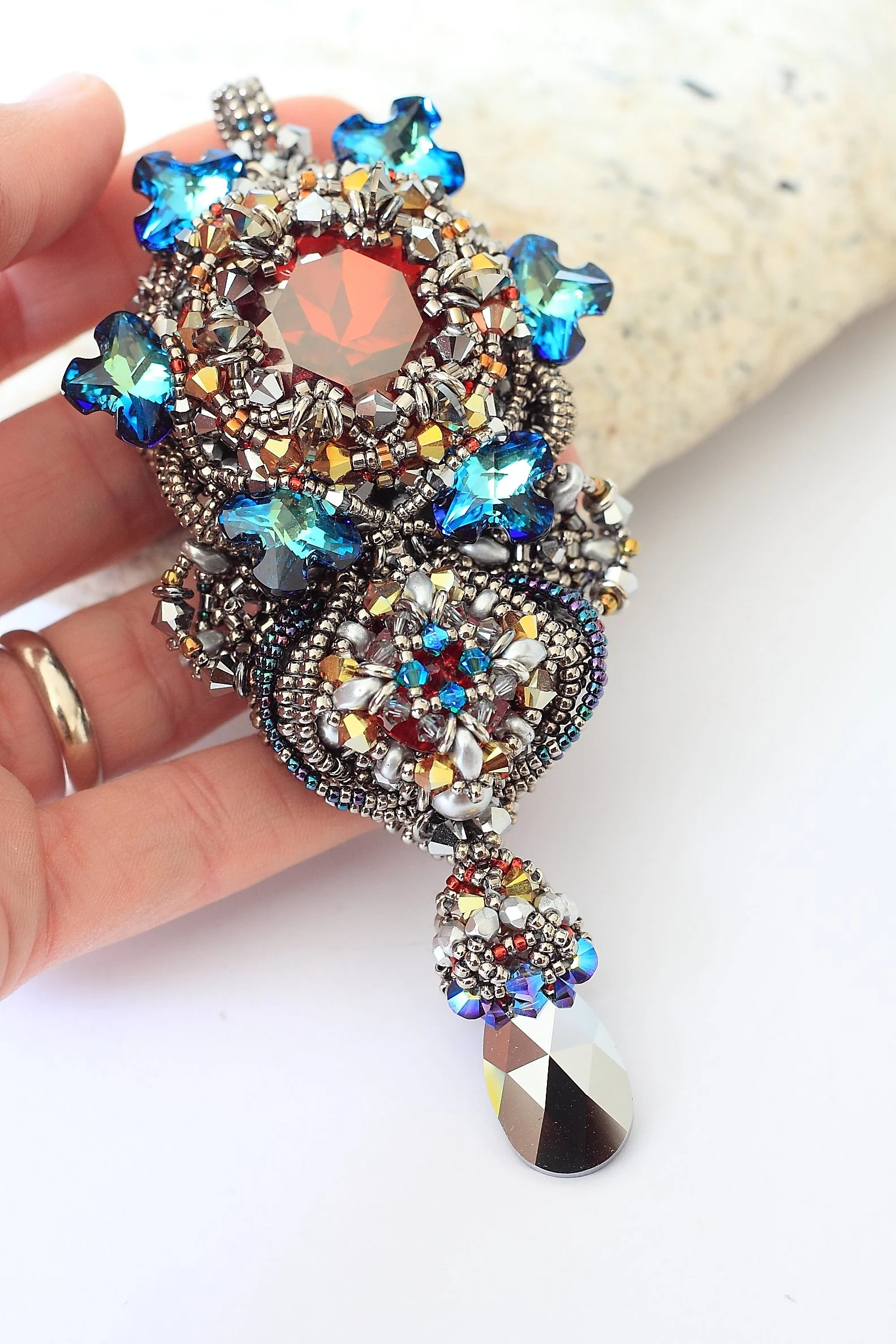Close-up of an ornate, beaded, and jewel-encrusted jewelry piece with large, geometric, reflective silver and amber-colored pendant and smaller blue, silver, and red decorative elements, held by a hand against a light background.