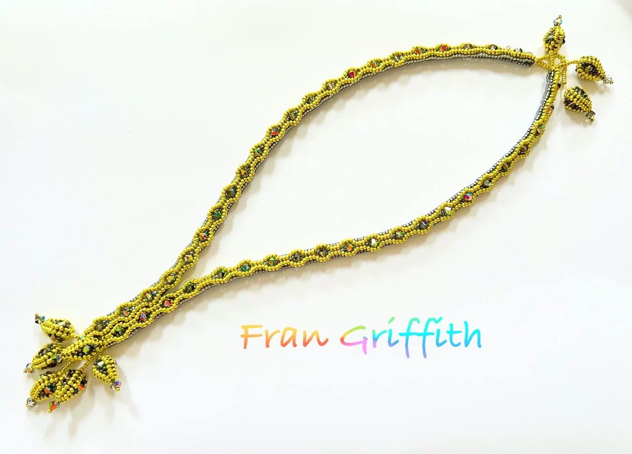 A colorful beaded necklace designed by Fran Griffith, with intricate yellow, green, black, and red beads, and small dangling bead ornaments at the end.