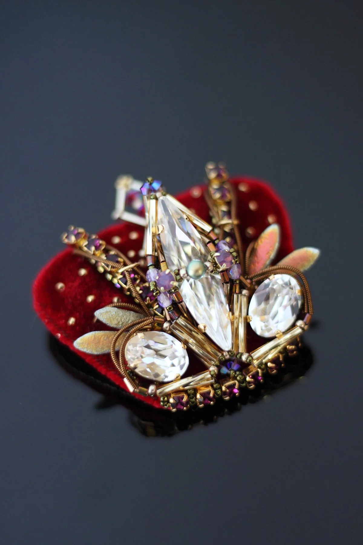 A jeweled brooch with a crown design, featuring large clear and purple gemstones, set on a red velvet backing.