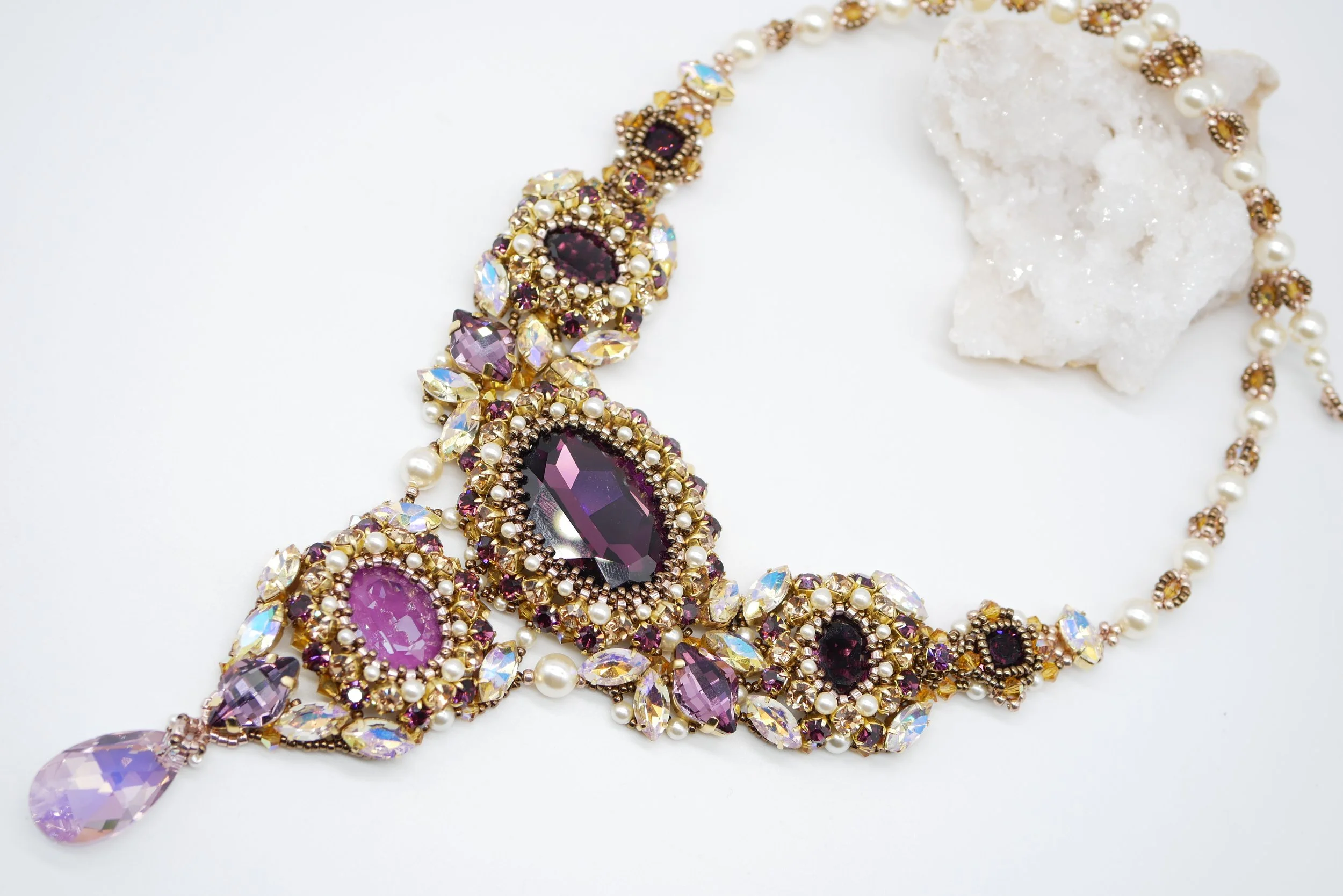 Close-up view of a purple gemstone necklace with gold, pearl, and iridescent accents, displayed on a white surface.