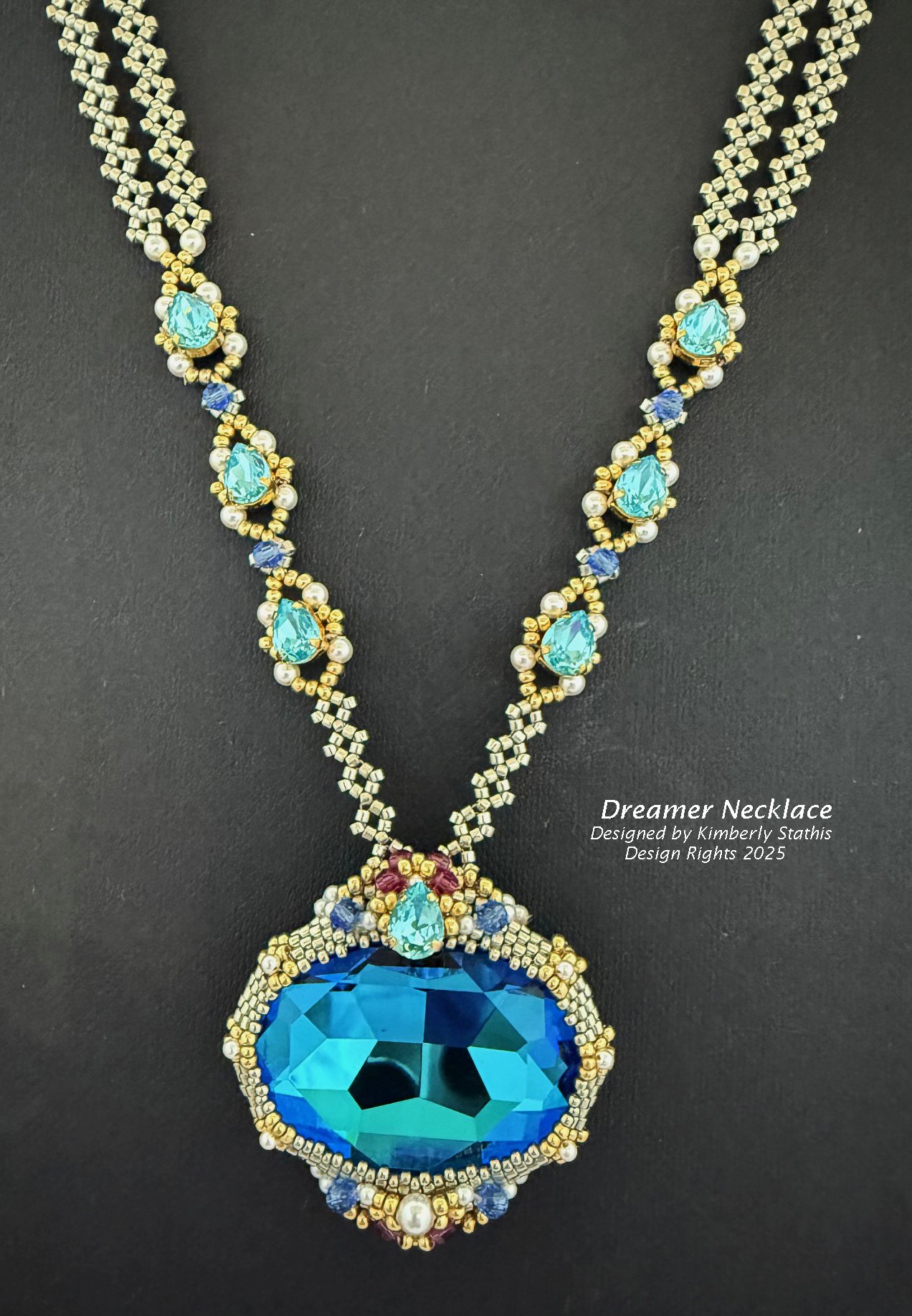 A beaded necklace named 'Dreamer Necklace' featuring a large blue gemstone at the center, surrounded by smaller blue, gold, and pearl beads. The necklace has an intricate design with multiple blue gemstones along its length, designed by Kimberly Stat