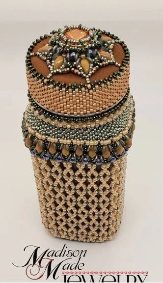 A cylindrical jewelry box decorated with intricate beadwork and embellishments, topped with a large ornamental brooch featuring orange, green, and black beads, and a large central gemstone.