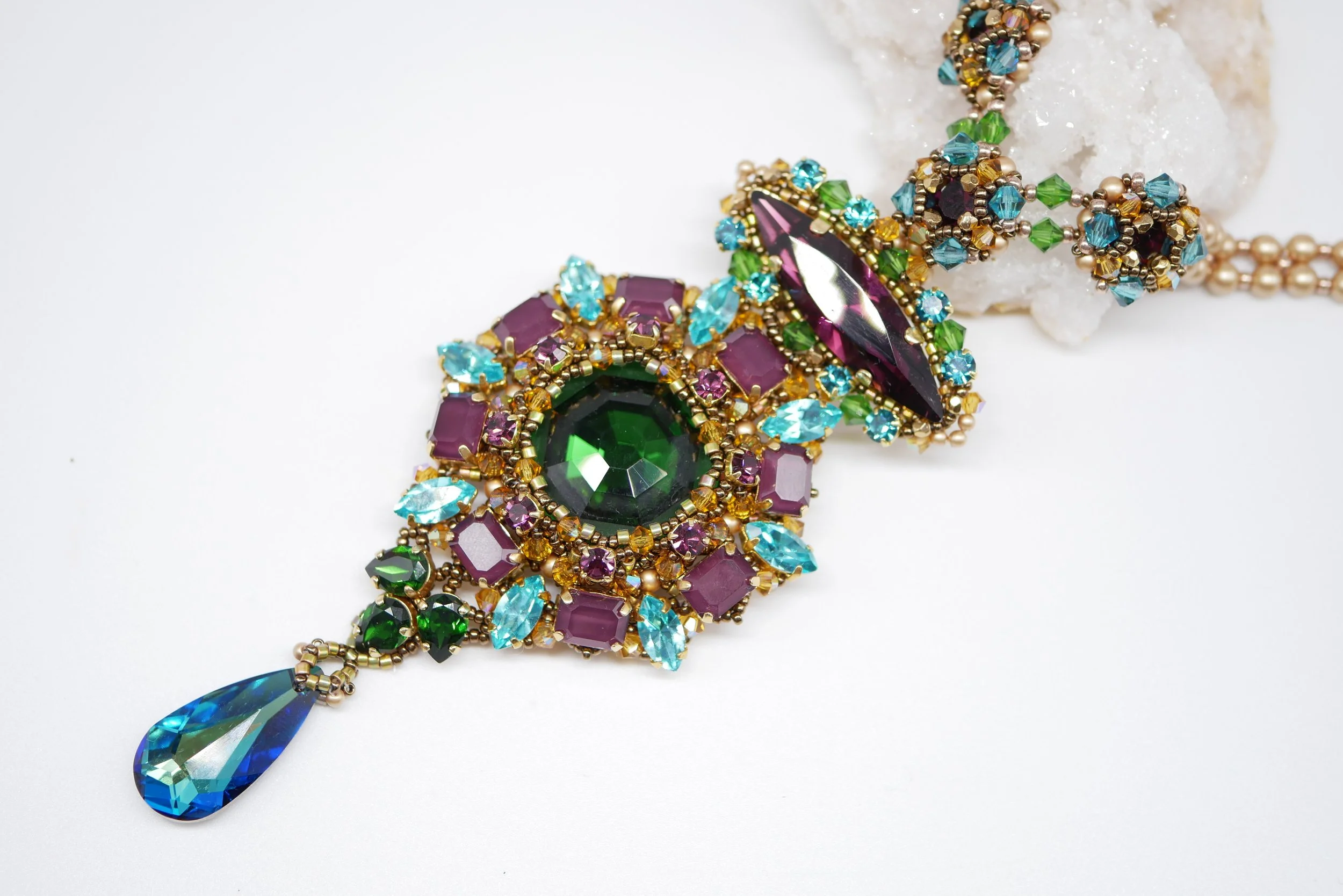 Colorful beaded jewelry with purple, green, blue, and gold accents, featuring a large teardrop-shaped blue pendant.