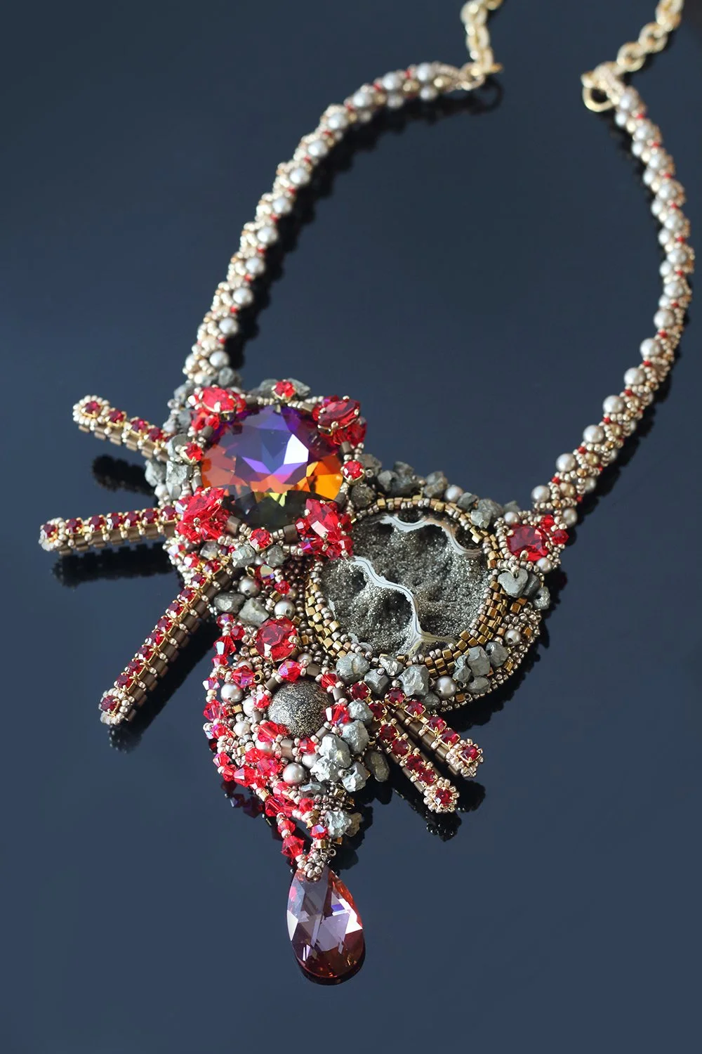 Close-up of a statement necklace with various beads, a large purple gemstone, and red accents, featuring a dangling teardrop-shaped gemstone on a black background.