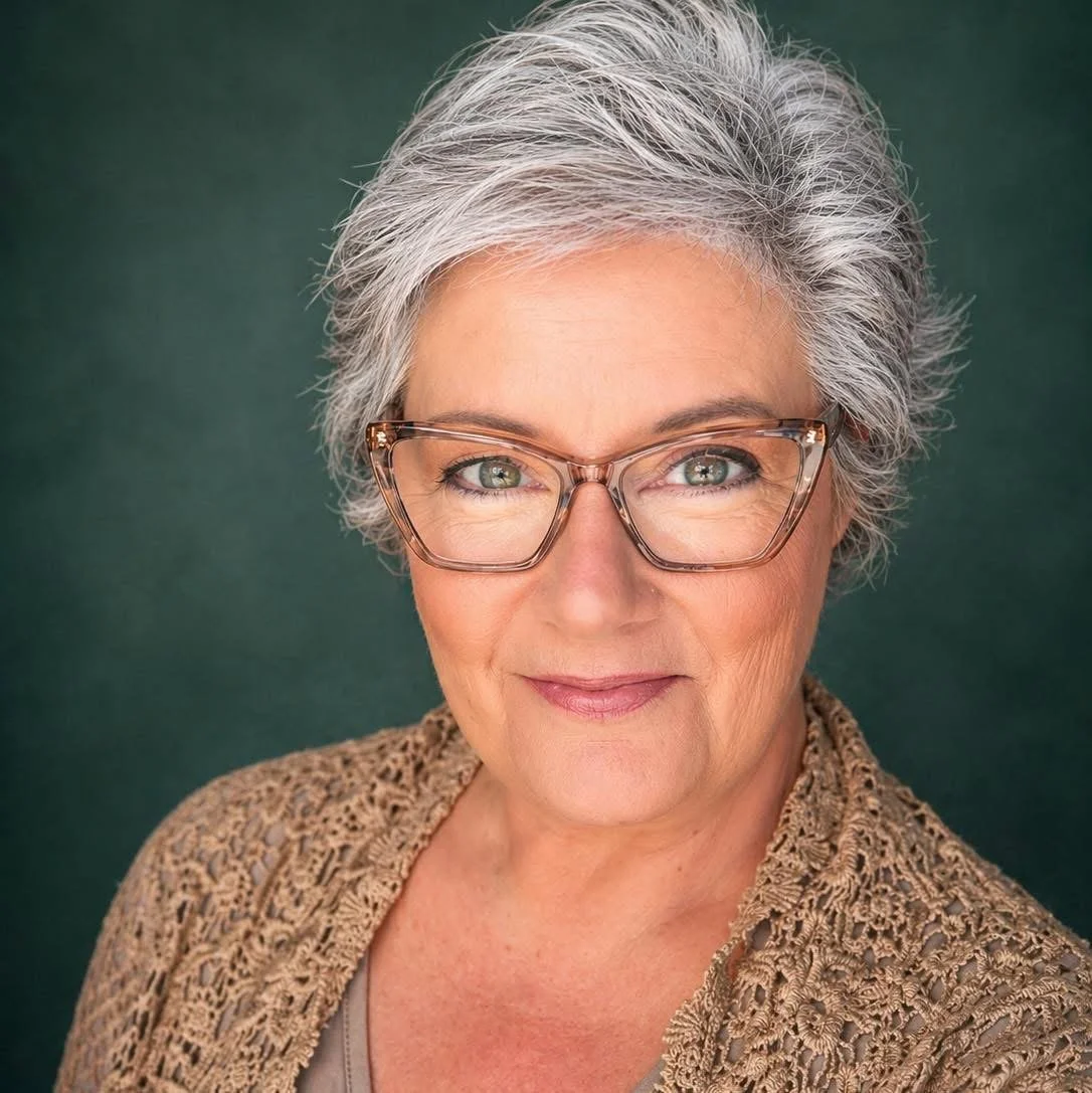 A portrait of an older woman with short gray hair, wearing glasses, a beige textured jacket, and a light shirt, smiling softly against a dark green background.