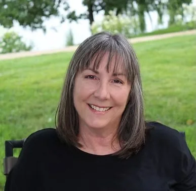 A woman with shoulder-length brown hair and bangs smiling outdoors on a grassy hill.
