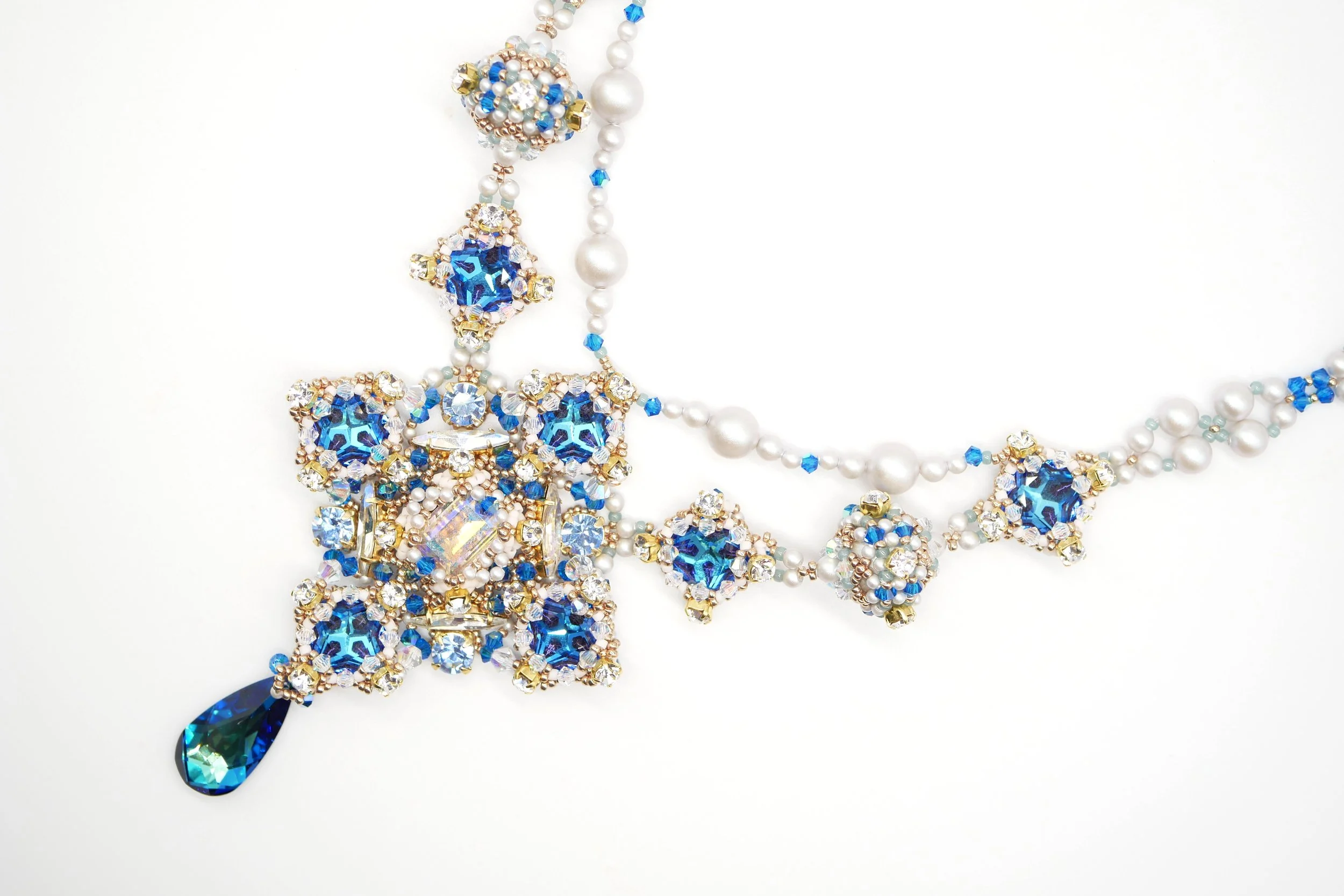 A necklace made of pearls, blue and clear crystals, and gold accents, featuring a large teardrop-shaped blue pendant.