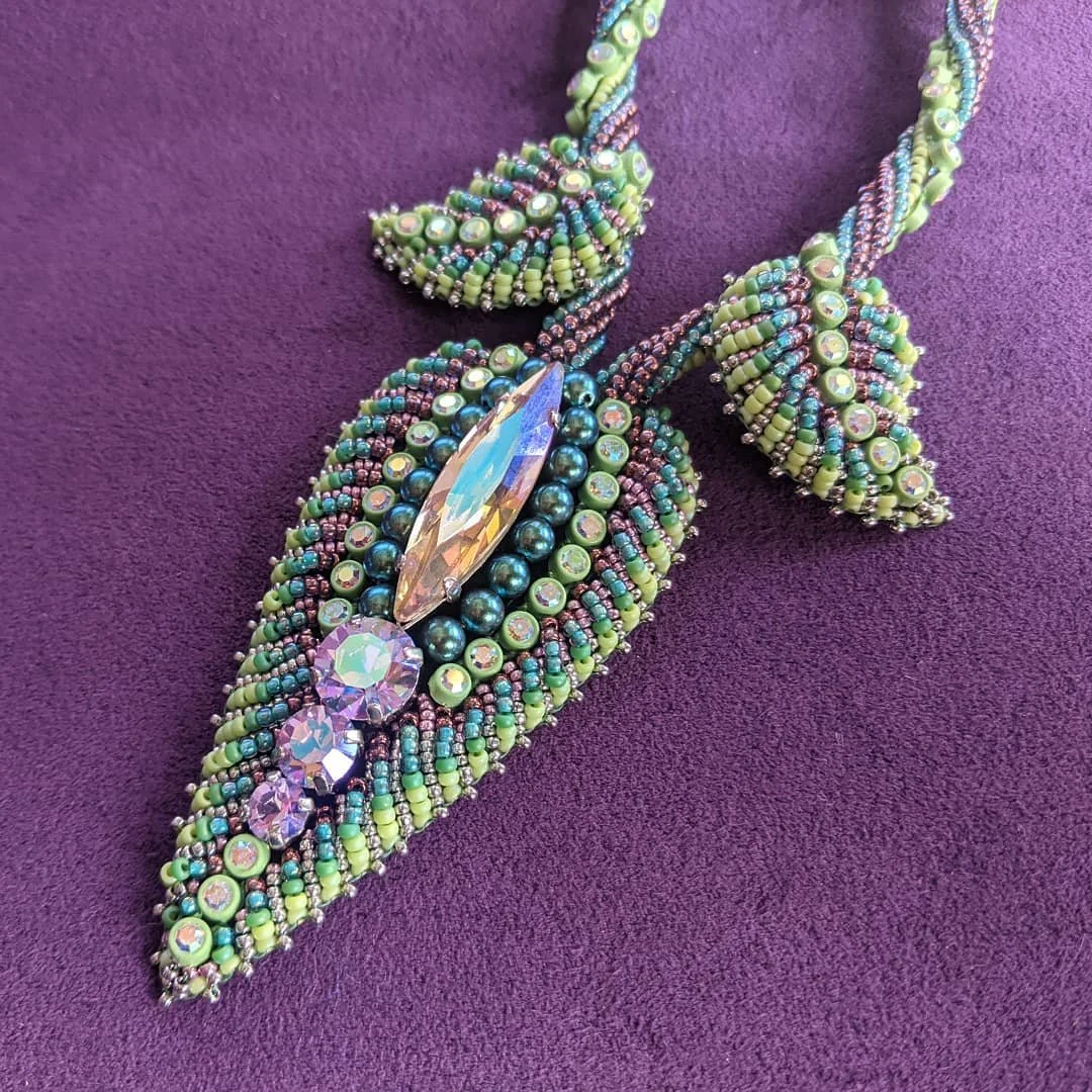 A beaded necklace with green, purple, and blue beads, featuring a large clear gem pendant and three smaller purple gems, resting on a purple fabric background.