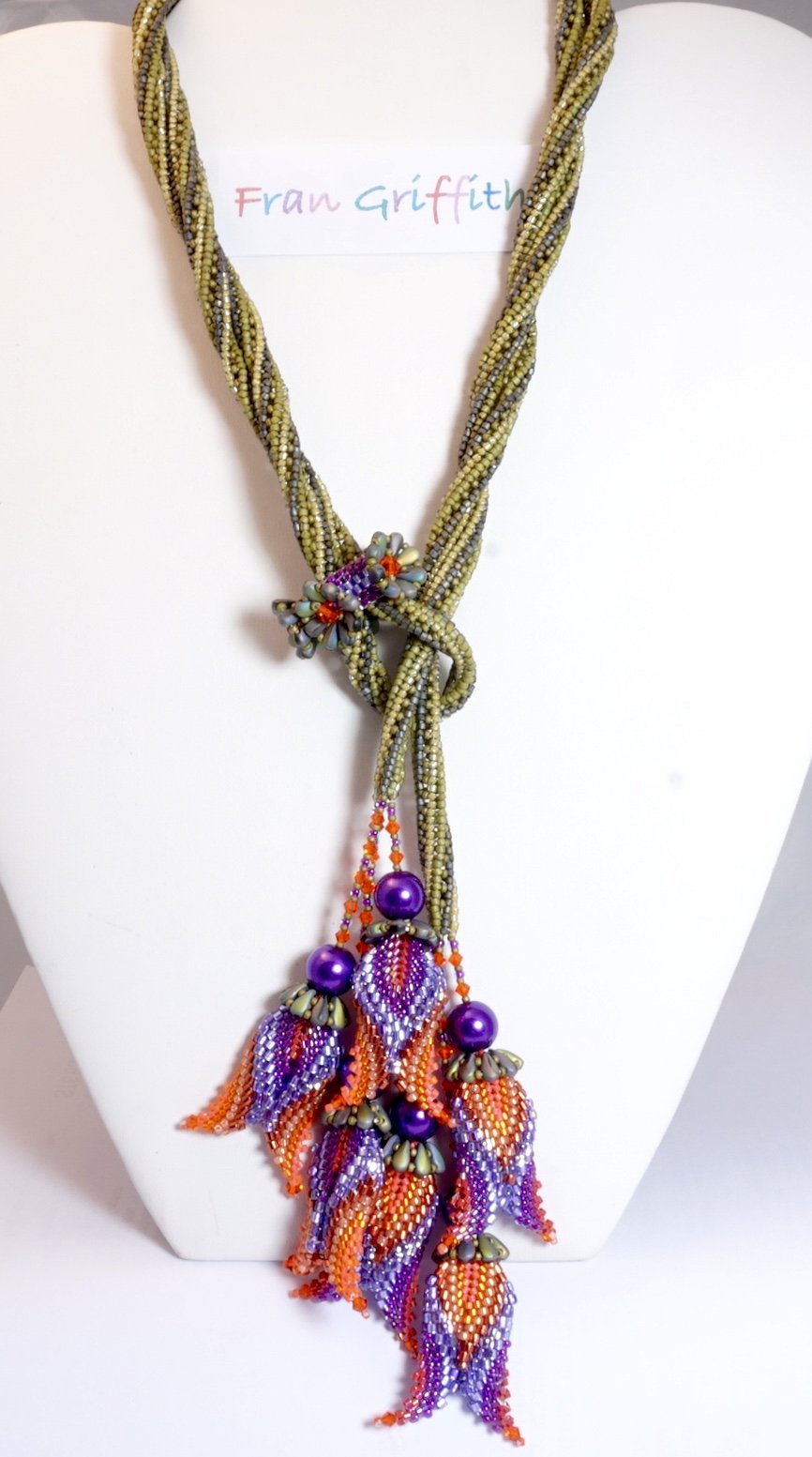 Colorful beaded necklace with purple, orange, and green beads, featuring a floral design and leaf-shaped pendants, displayed on a white bust.