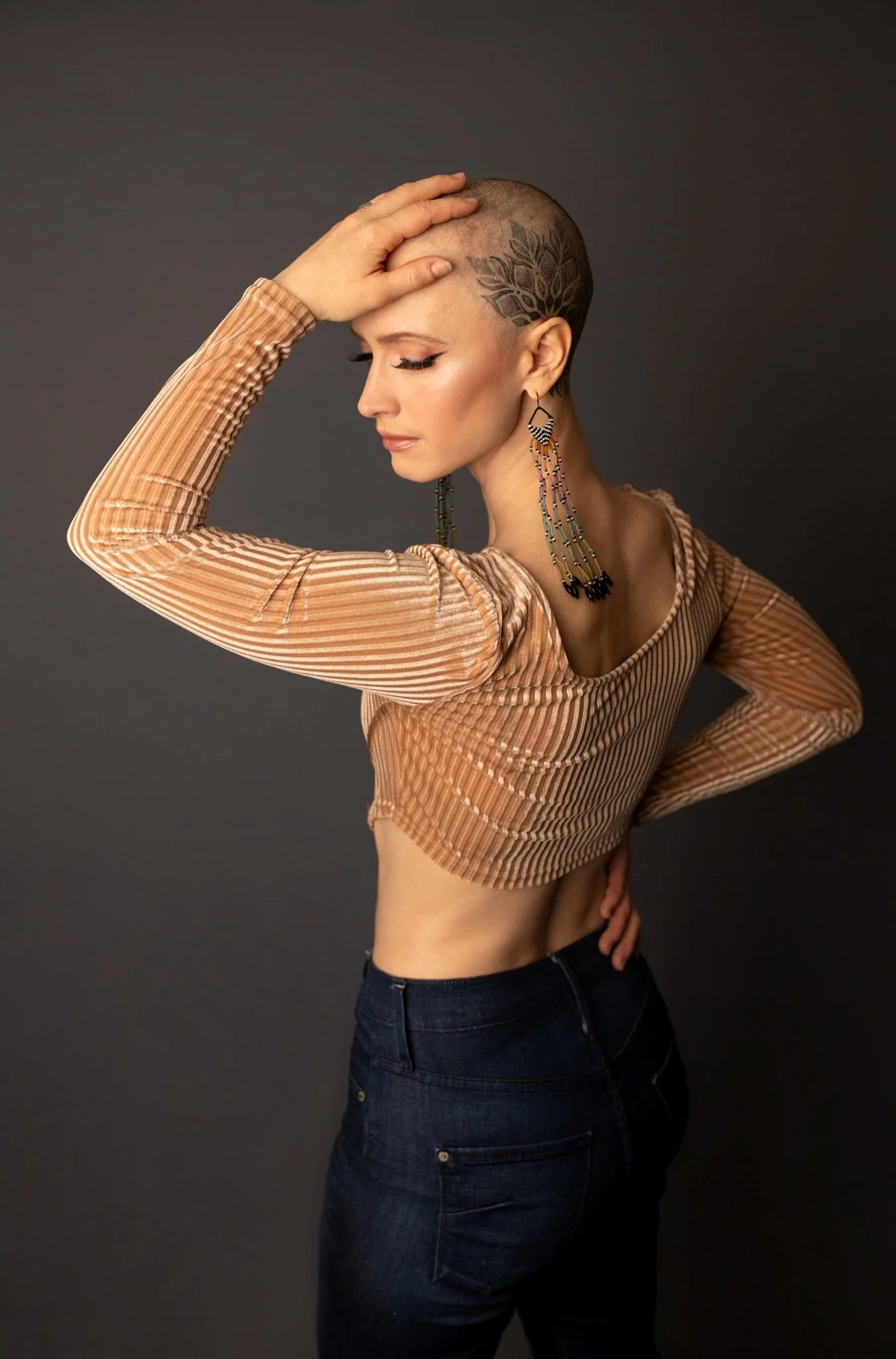 A woman with a shaved head and a leaf tattoo on her scalp, wearing a striped long-sleeve top and high-waisted jeans, standing against a dark background, with her eyes closed and one hand on her head.