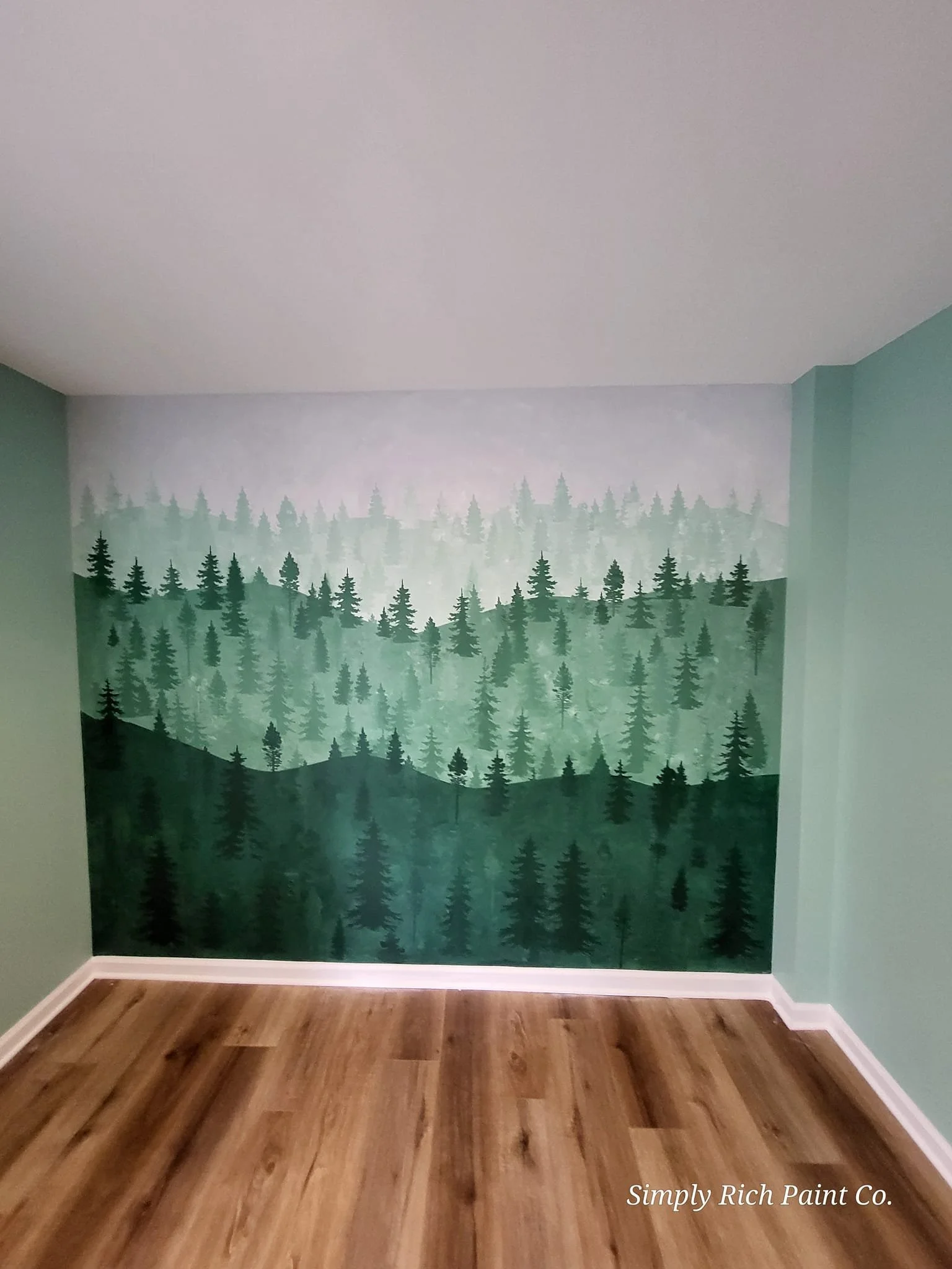 Room with a forest-themed mural featuring layers of green pine trees painted on the wall, with hardwood flooring and baseboards.
