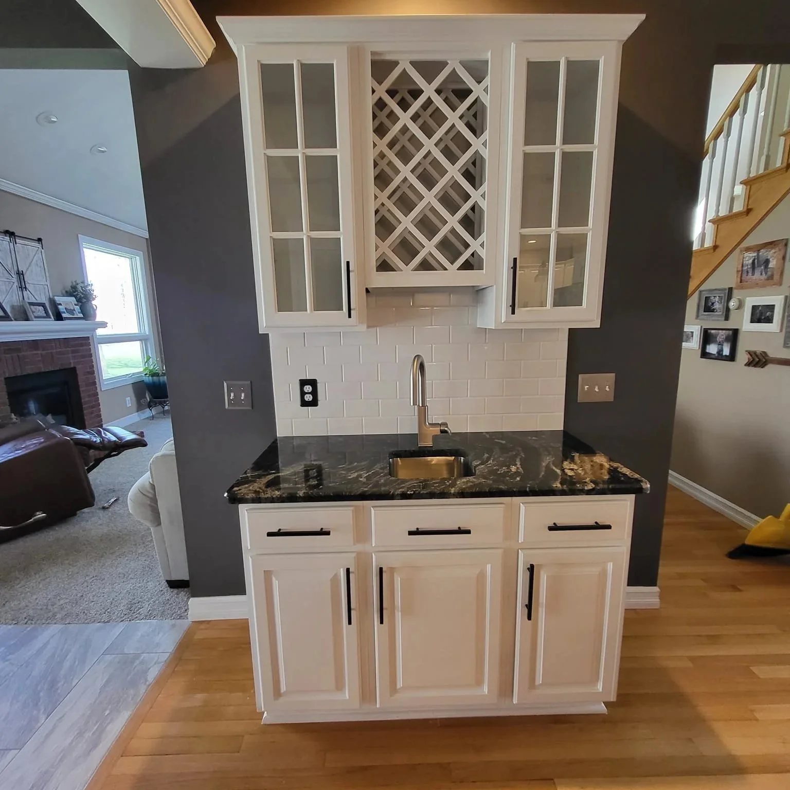 Kitchen mini bar with a black marble countertop, white cabinets, a gold sink, and a wine rack above. Open living room with a fireplace in the background.