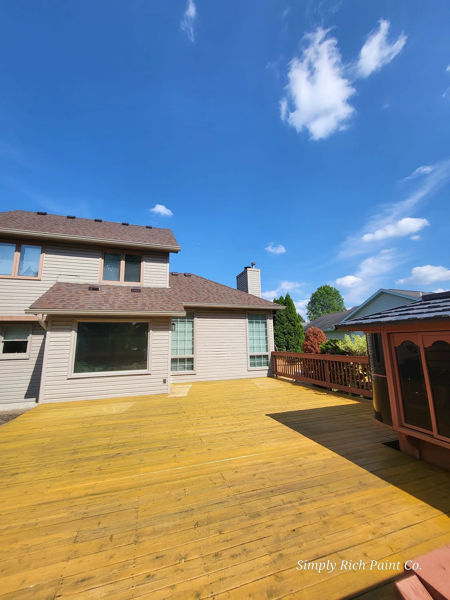 A backyard wooden deck with a house featuring beige siding, large windows, and a chimney under a blue sky with scattered clouds.