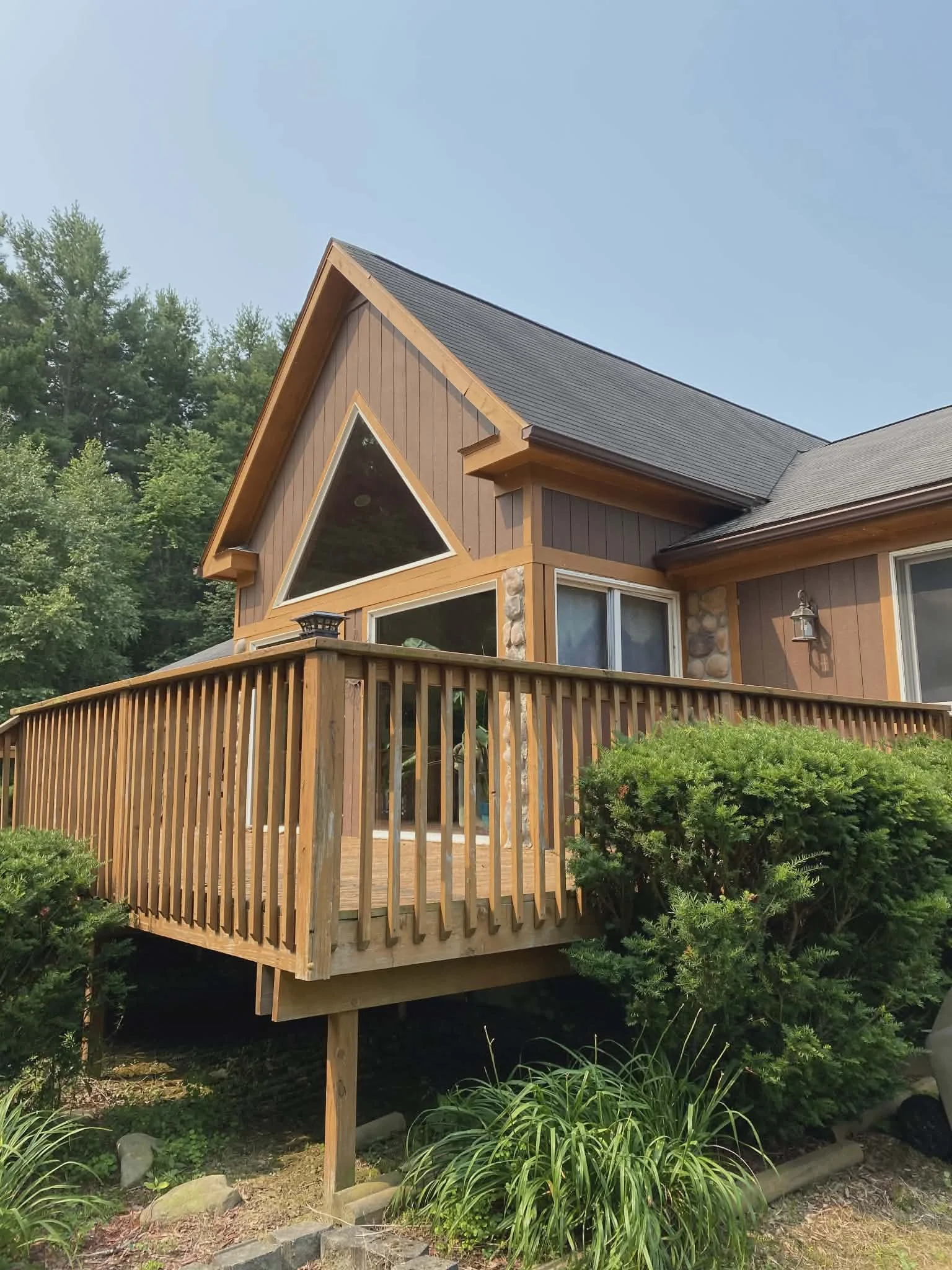Photo of a house with a wooden deck and large triangular window, surrounded by greenery and bushes.
