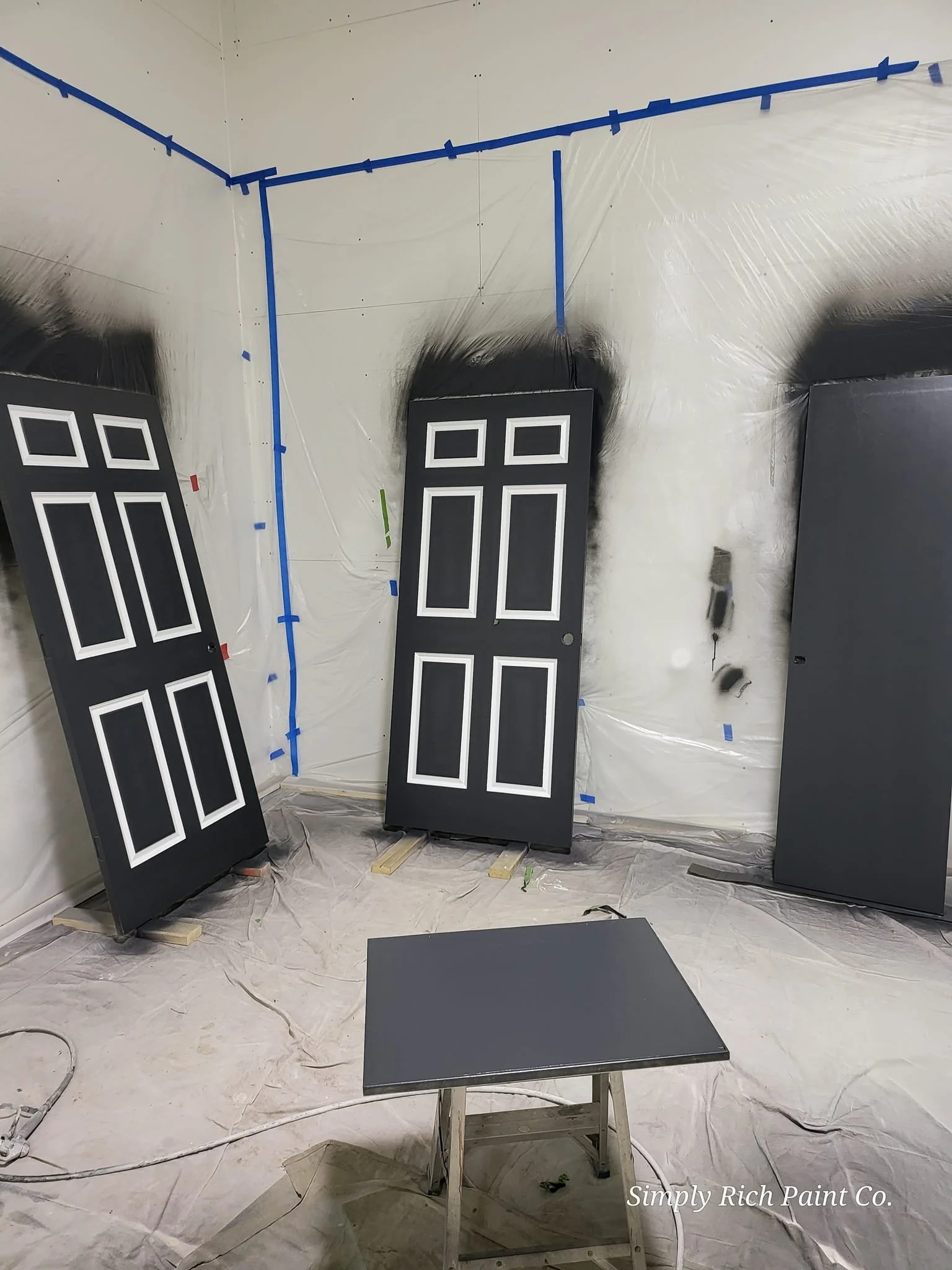 Three black painted doors leaning against a wall, with a small black table in front. The room is covered with plastic sheeting, and the edges around the doors are spray painted black. Blue painter's tape is visible on the corners and ceiling.