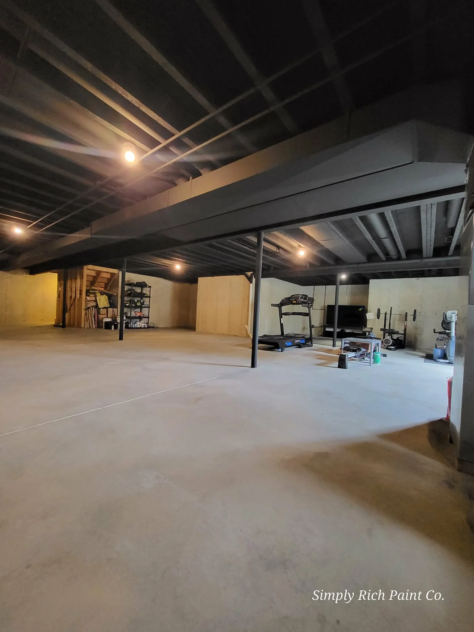 An unfinished basement with concrete floor, black ceiling, and a small workout area with a treadmill and some weights, illuminated by hanging lights.