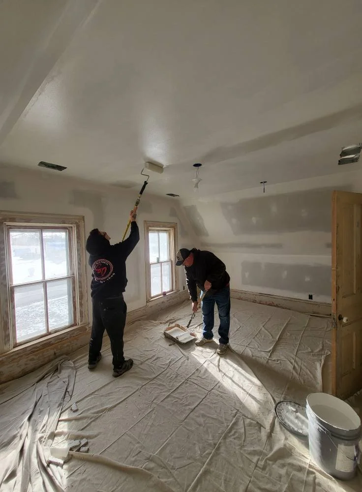 Two people painting the ceiling of a room with a roller brush, while another person paints near the wall on the floor, with windows and covered floors.