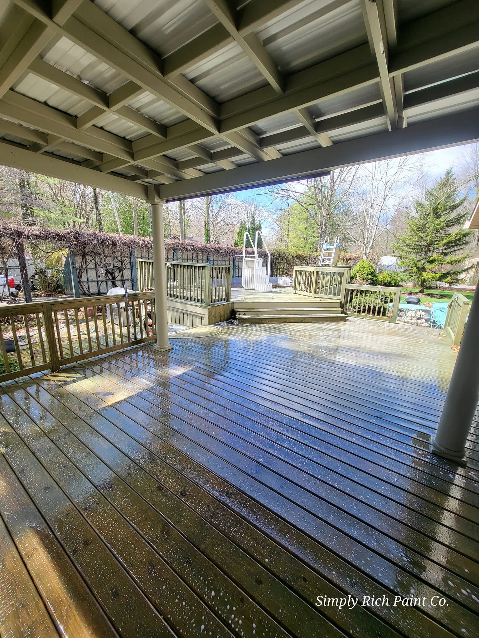 A wet wooden deck under a covered patio, overlooking a backyard with trees, a white ladder, and outdoor furniture.