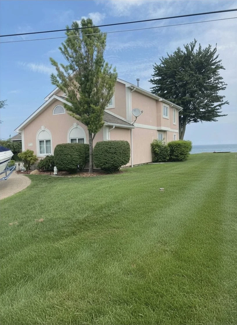 A two-story peach-colored house with white trim, surrounded by green lawn and bushes, with two trees near the house and a view of water in the background. The sky is blue with a few clouds.